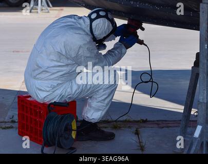 Worker in protective suit sands the hull of a yacht while sitting on a red crate, with tools and electrical cords visible on the ground Stock Photo