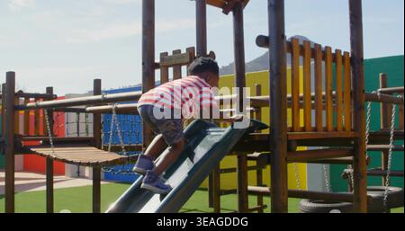 Climbing child wearing striped tee camo shorts on plastic slide at playset, with rope bridge Stock Photo