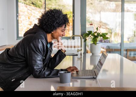 African American man leaning on kitchen island, typing on silver laptop, wearing leather jacket Stock Photo