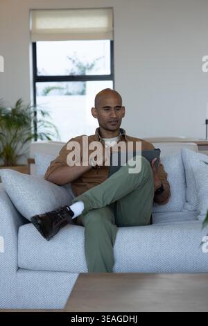 Middle-aged Indian man sitting cross-legged on light grey sofa holding tablet in living room Stock Photo