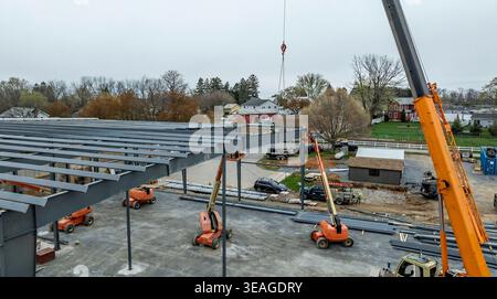 Workers are using cranes and lifts to build a new structure. Various machines are positioned on a construction site. Nearby houses are visible in the background under cloudy skies. Stock Photo