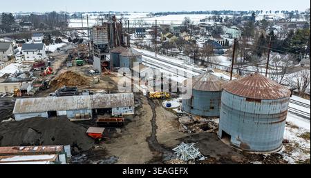 Workers are present at a construction site with silos and old structures. The area shows piles of dirt, building materials, and a nearby train track. Snow covers some parts of the ground. Stock Photo