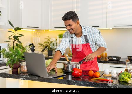 Indian man cooking healthy soup while smiling at laptop in home kitchen for weight loss meal prep. Arabian guy following online recipe for clean eating lunch with focus joy comfort during work break. Stock Photo