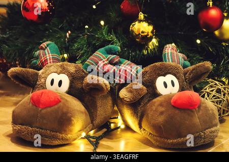 Cozy plush reindeer slippers by a decorated christmas tree with warm fairy lights and red-gold ornaments, festive holiday home scene. Stock Photo