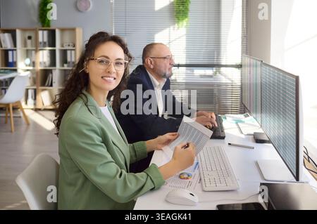 Smiling analyst with report beside colleague working on spreadsheets at desktop Stock Photo
