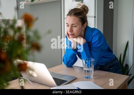 Thoughtful mature woman engaged in self learning process online on laptop at home in kitchen. Stock Photo