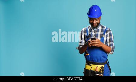 Construction worker texting messages on smartphone app, using social media browser in studio. Young craftsman browsing online website, wearing building overalls and safety helmet. Stock Photo
