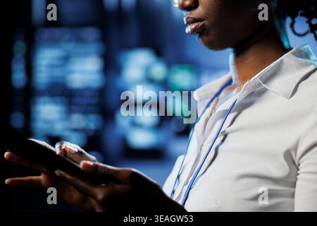 Focused admin in data center uses tablet to prevent server machines overload. Expert between server mainframes using device to ensure enough network bandwidth for smooth operations Stock Photo