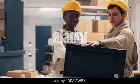 Diverse women planning shipment talking about order in storage room space, checking products logistics on list in packages. Supervisor and employee working in team, delivery service stock. Stock Photo
