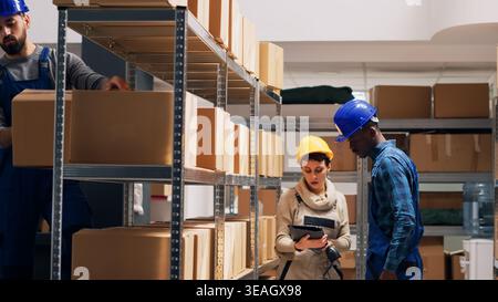 Diverse depot workers using digital tablet to scan goods, pointing scanner at barcodes on boxes to check stock logistics. Young employees in overalls working with cargo inventory. Stock Photo