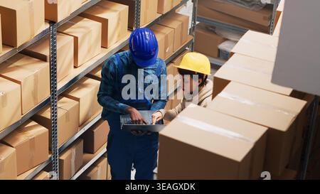 Multiethnic team of people checking stock list on laptop, using logistics to count products on shelves. Two employees working in storage room depot, planning order distribution. Handheld shot. Stock Photo