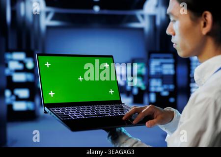 IT consultant with green screen laptop between server hub clusters providing processing resources for businesses worldwide. Worker uses mockup device to fix data center mainframes Stock Photo