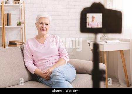 Friendly senior woman vlogger sitting on sofa dating online Stock Photo