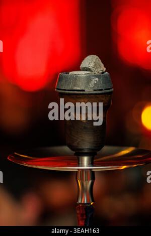 A close-up of a traditional hookah head featuring glowing coals, set against a blurred red bokeh background. Stock Photo