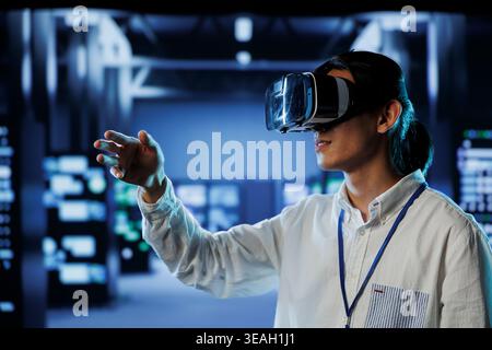 Qualified programmer in data center uses virtual reality to prevent system overload. Specialist with VR goggles in server hub ensuring enough network bandwidth for smooth operations Stock Photo