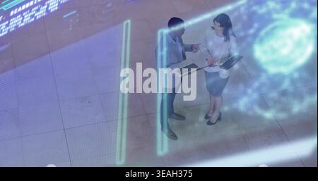 Discussing two colleagues in suits reviewing documents in lobby with laptop, tablet, hologram Stock Photo