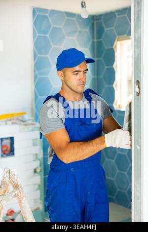 Industrial worker, handyman in blue construction overalls installing tiles on bathroom walls Stock Photo