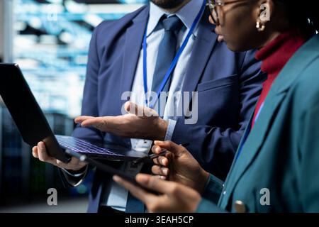 Close up of data center coworkers using laptop and tablet, evaluating infrastructure diagnostic reports. Server hub IT staff members working together to automate supercomputers processing Stock Photo