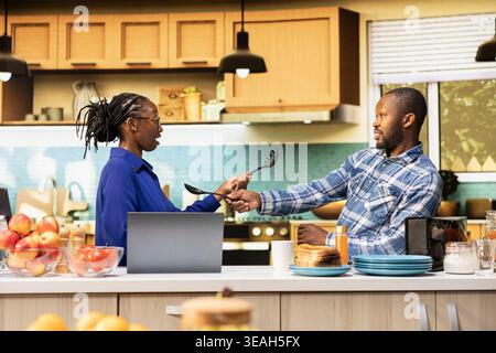Black young couple starting a duel with kitchen utensils for the game, playful fight with fake swords during the goofy war. Childish silly adversaries having fun together in the morning. Stock Photo