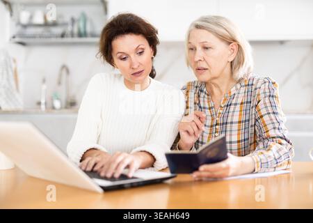 Old woman showing her passport to middle-aged woman sitting next to her and working on laptop Stock Photo