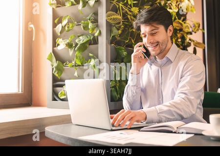 Cheerful businessman working on laptop and talking on phone in cafe Stock Photo