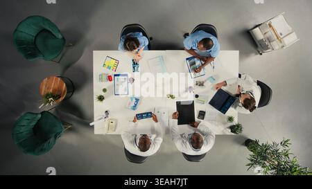 Top down view of specialists and medics collaborating in a hospital conference room. Healthcare staff brainstorms treatment ideas and wellness through teamwork and communication. Stock Photo
