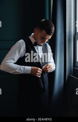 Groom Buttoning Vest Near Window, Preparing For Ceremony In Moody Room With Deep Teal Wall And Heavy Curtain. Focused Expression, Crisp White Shirt Stock Photo