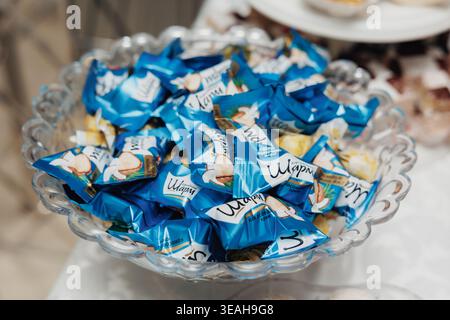 Glass Bowl Filled With Wrapped Candies In Blue Packaging, Glossy Wrappers Catching Light, Closeup Shot On Party Table With Blurred Desserts And Plates Stock Photo
