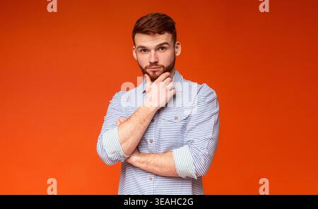 Man in Striped Shirt Poses Against Orange Background With Thoughtful Expression Stock Photo