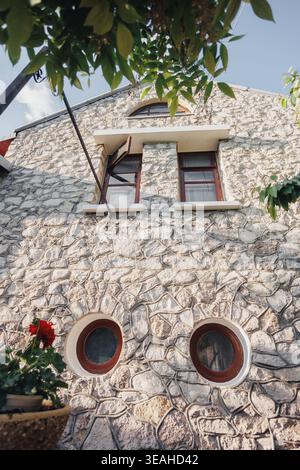 Sunlit Stone Facade With Round Windows, Arched Upper Window Framed In Wood, Terracotta Planter Brimming With Geranium, Climbing Vines And Leafy Canopy Stock Photo