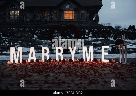 Dusk Proposal Scene With Illuminated MARRY ME Letters, Rose Petals Scattered Across Wooden Shore, Glowing Lantern, Distant House Window Casting Warm Stock Photo