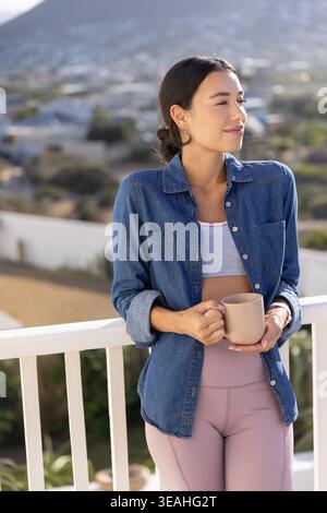 Female standing on white balcony railing holding beige ceramic mug gazing at houses and mountain Stock Photo