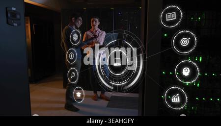 Discussing IT staff woman in button-up, man in sweater, holding tablet near hologram UI and servers Stock Photo