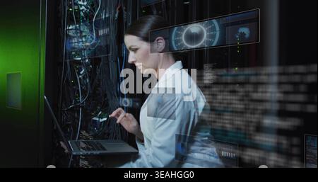 Working IT technician in white labcoat typing laptop handling cables in server room, holographic UI Stock Photo