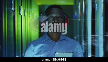 Datacenter tech in blue shirt monitoring racks holding tablet, wearing red earmuffs HUD visible Stock Photo