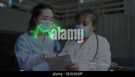 Studying two masked clinicians in scrubs, lab coat with stethoscope reviewing tablet, ECG overlay Stock Photo