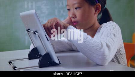 Studying Chinese girl using tablet at school desk, wearing sweater-headband near chalkboard Stock Photo