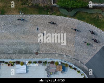 Aerial view of a group of cyclists riding on a curved cobblestone path next to a building terrace with outdoor seating, potted plants and green bushes Stock Photo