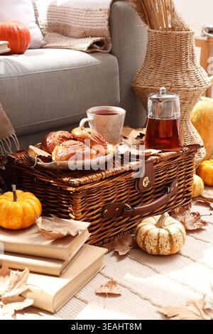 Autumn composition with teapot, cup of hot beverage, tasty buns and pumpkins on wicker basket in living room Stock Photo