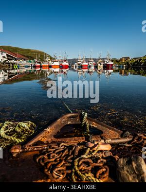 A weathered old wooden rowing boat resting on the shore in the foreground of a calm Norwegian harbour with fishing boats and reflections behind. Stock Photo