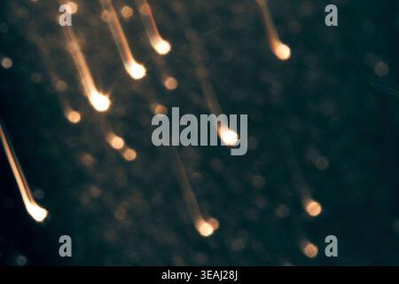 Golden Light Streaks Descending In Blurred Motion Against Dark Speckled Background With Bokeh Highlights In Low Light Abstract Setting. Stock Photo