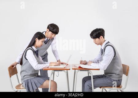 A group of high school students in uniform work together on a project, sitting at desks with laptops and books, in a modern classroom. Stock Photo