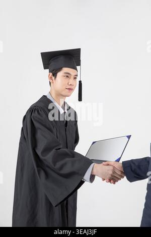 A young Asian man in a graduation gown shakes hands with an adult, holding a diploma in front of a white background. Stock Photo