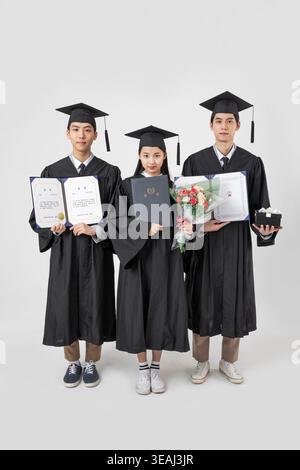 Three young Asian graduates, holding their certificates and gifts, stand together in their caps and gowns. Stock Photo