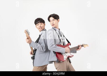 Two Asian high school students, both wearing school uniforms, playfully strum and smile at each other while holding their guitars. Stock Photo