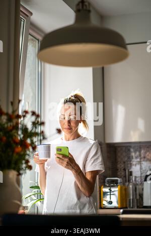 Relaxed thoughtful woman drinking coffee scrolling smartphone at cozy sunny kitchen beside window. Stock Photo