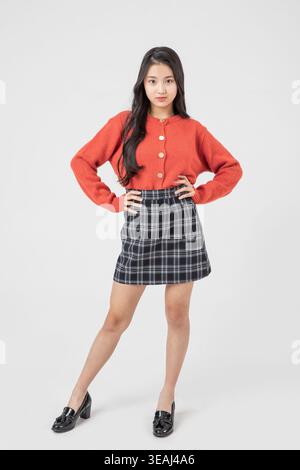 A young Asian woman stands confidently with her hands on her hips, wearing a red sweater and a plaid skirt, set against a plain white background. Stock Photo