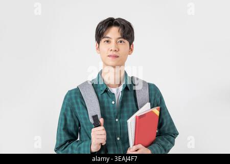 A young Asian student holds a stack of books and a backpack, standing in a neutral studio setting. Stock Photo