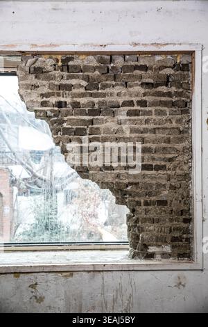 A crumbling brick wall with a large, jagged hole in it, exposed to a winter landscape through a broken window. Stock Photo