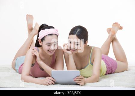 Two young women lying on a white rug, laughing while looking at a tablet together. Stock Photo